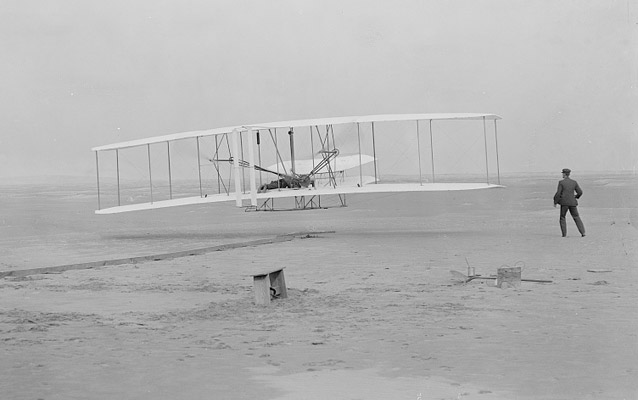 ภาพการบินครั้งแรกของ Wright Flyer ปี 1903 ออร์วิลล์อยู่บนเครื่องและวิลเบอร์อยู่ข้างเครื่อง